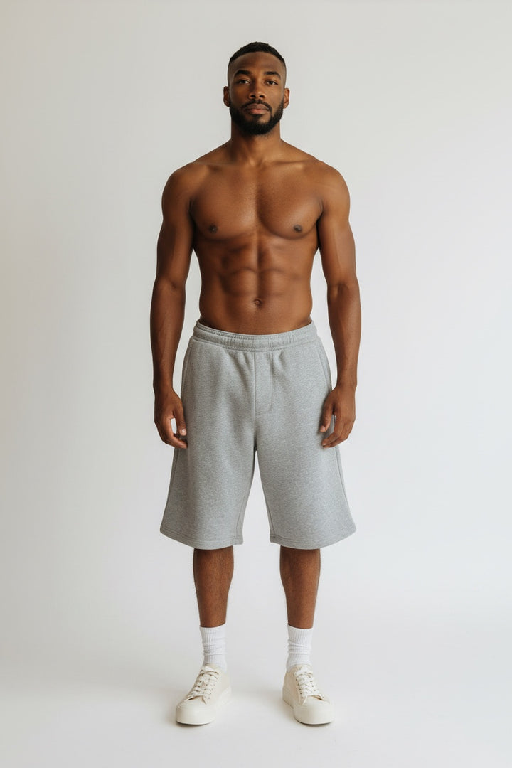 A shirtless man stands against a light gray studio background wearing light gray heather sweat shorts, white socks, and white sneakers.