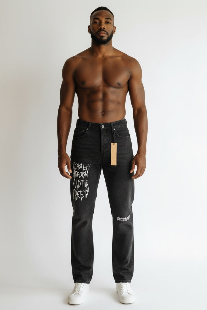 A shirtless man modeling black distressed jeans with white graphic text on the left leg, standing against a plain light gray studio background.