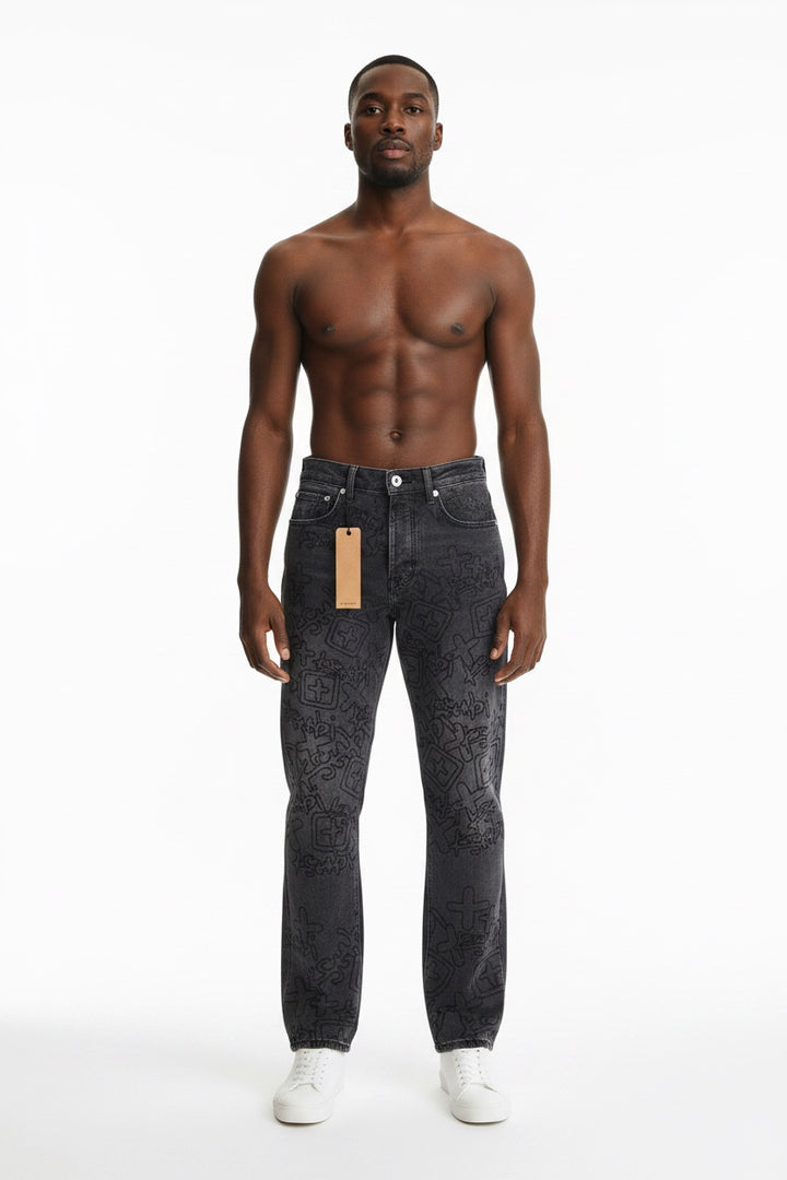 A shirtless male model stands against a plain white studio background wearing dark gray patterned jeans with a visible paper hangtag and white sneakers.